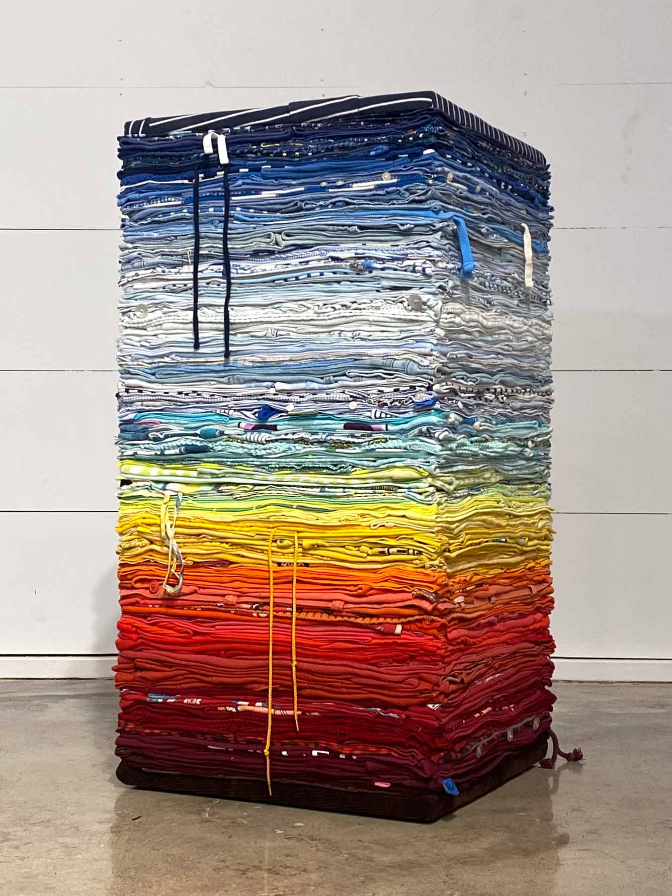Table made out of layers of used clothing, arranged in a gradient from maroon red, orange, yellow, sky blue, to navy blue. Clothing transformation into a functional soft abstract sculpture with sustainability in mind as core value to push against mindless consumer culture and waste. The table is a column shape. Made by Derick Melander.